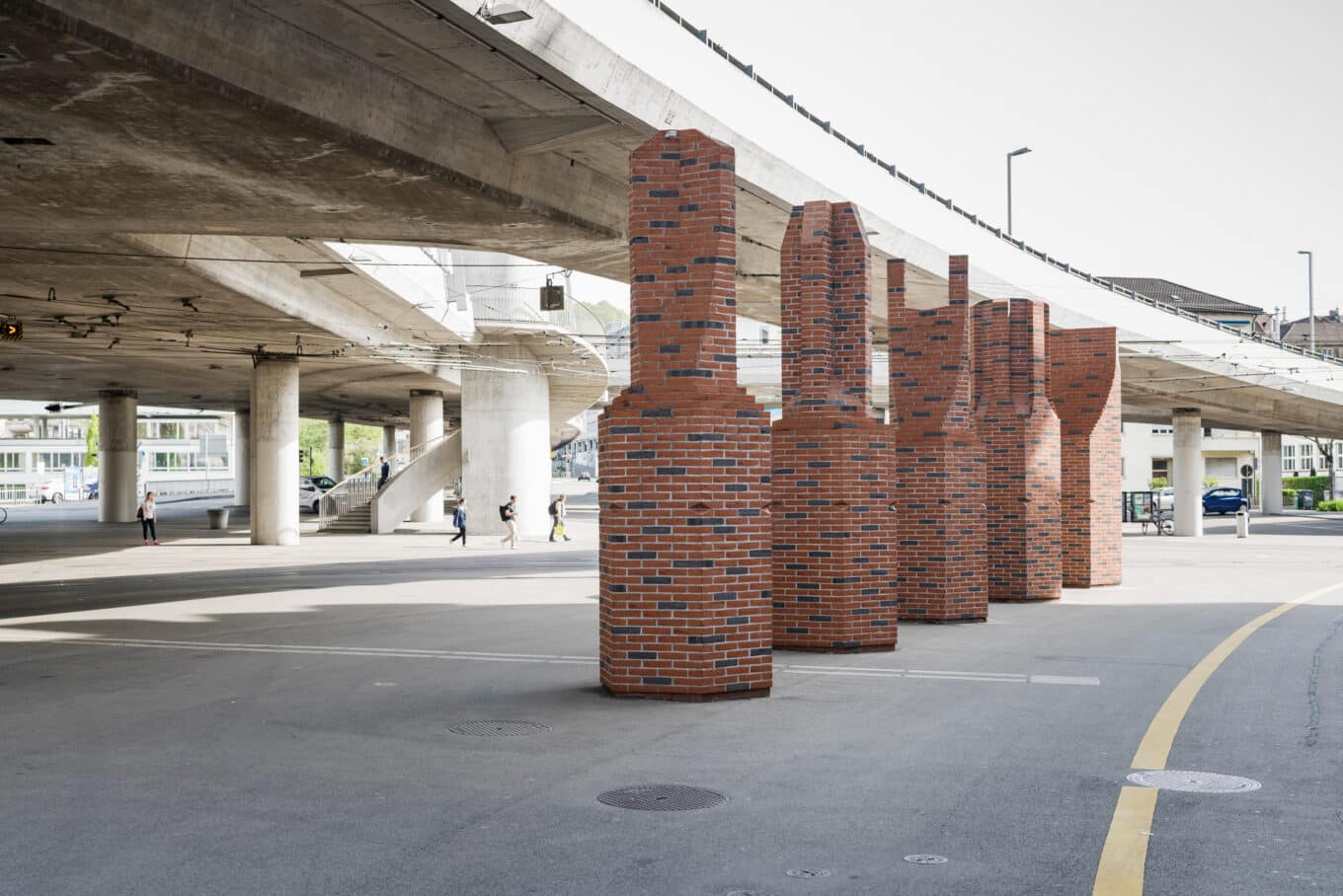 Fünf kaminähnliche Ziegelsäulen in einer Reihe unterhalb einer erhöhten Betonkonstruktion mit sichtbaren Gehwegen und vereinzelten Fussgängern.