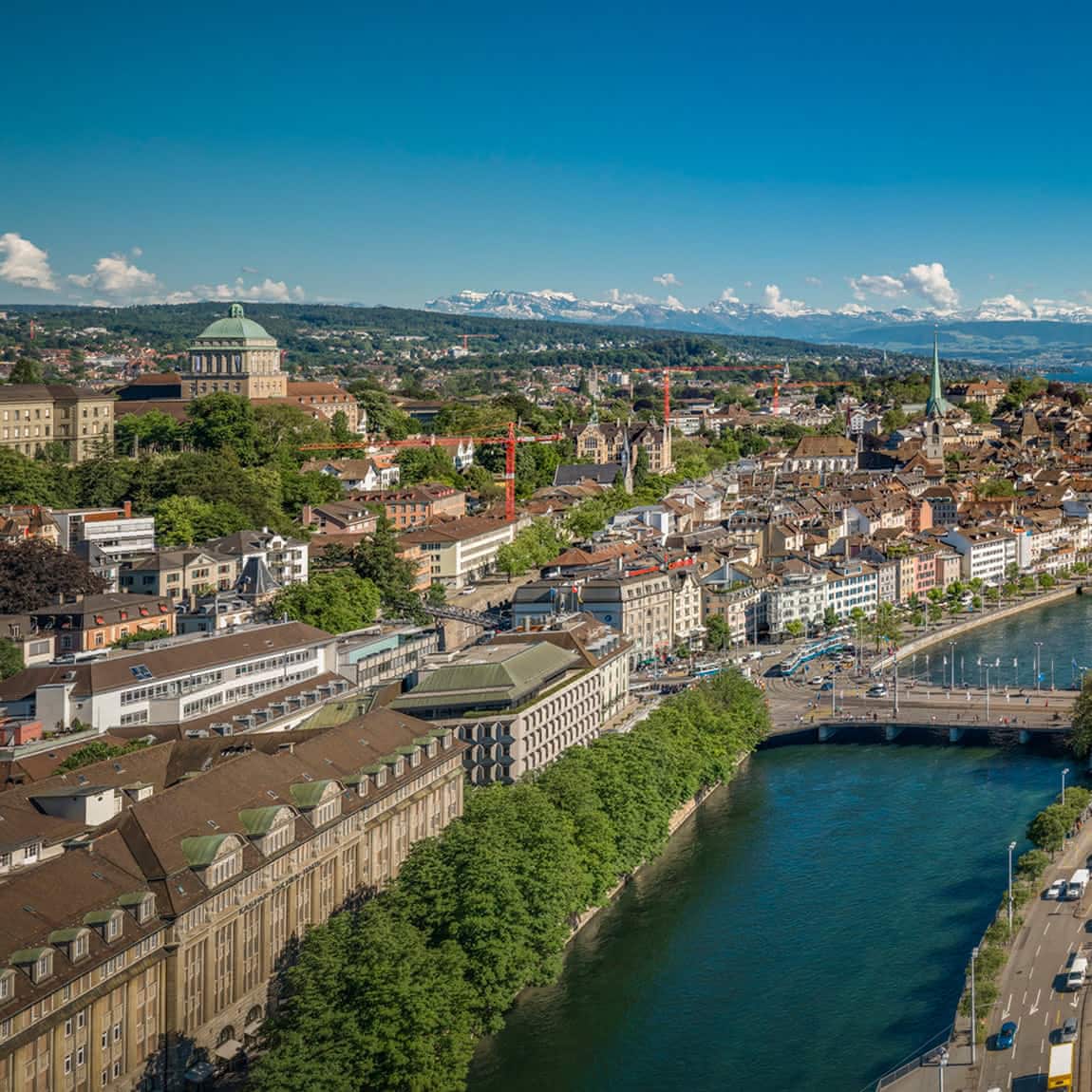 Luftaufnahme auf Zürich und die Limmat, umgeben von grünen Hügeln und Bergen in der Ferne unter einem klaren blauen Himmel.