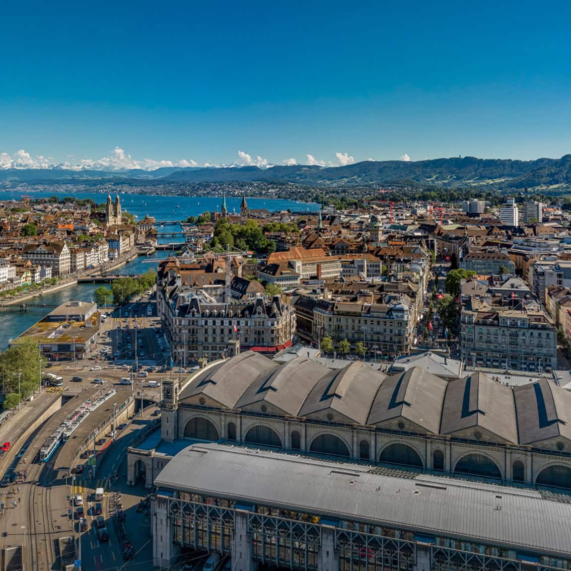 Luftaufnahme auf Zürich und die Limmat, umgeben von grünen Hügeln und Bergen in der Ferne unter einem klaren blauen Himmel.