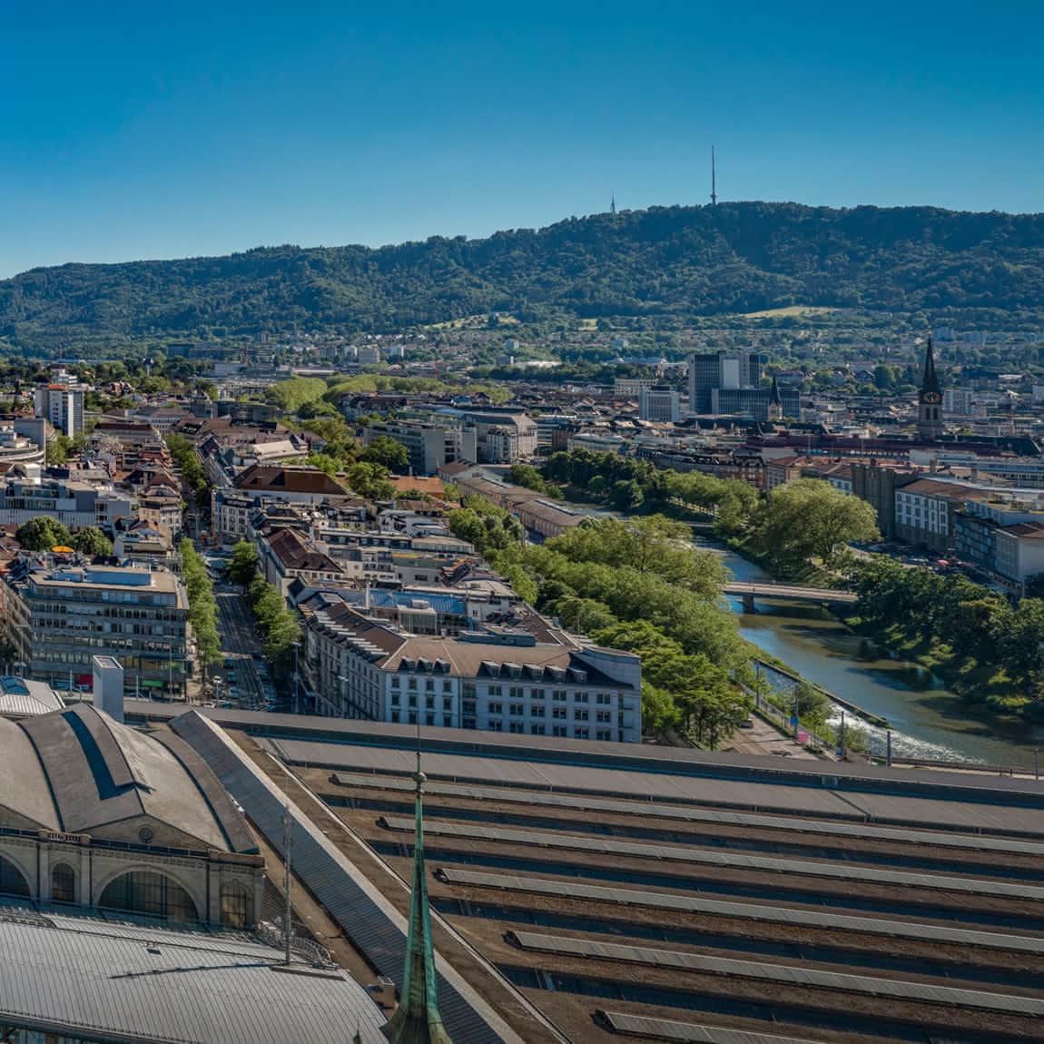 Luftaufnahme auf Zürich und die Limmat, umgeben von grünen Hügeln und Bergen in der Ferne unter einem klaren blauen Himmel.