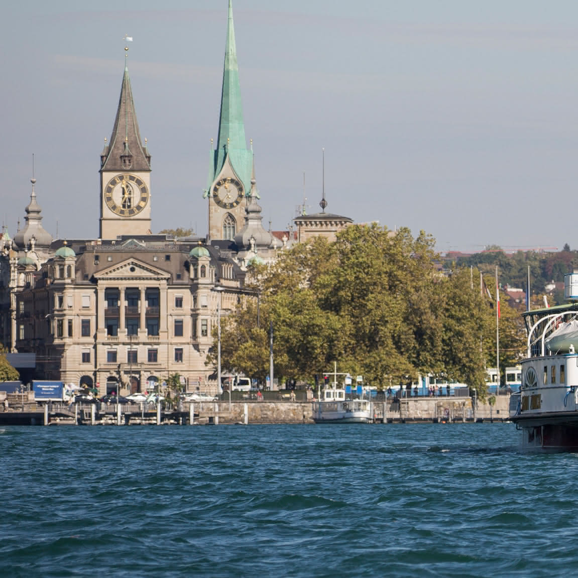 Blick auf die Uferpromenade vom Zürichsee mit historischen Gebäuden und Kirchtürmen mit Zifferblättern, umgeben von Bäumen.