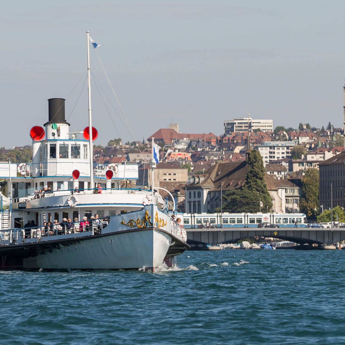 Ein grosses weisses Boot mit roten Akzenten liegt in der Nähe einer Stadtküste vor Anker. Im Hintergrund sind unter einem klaren Himmel Gebäude und eine Brücke zu sehen.