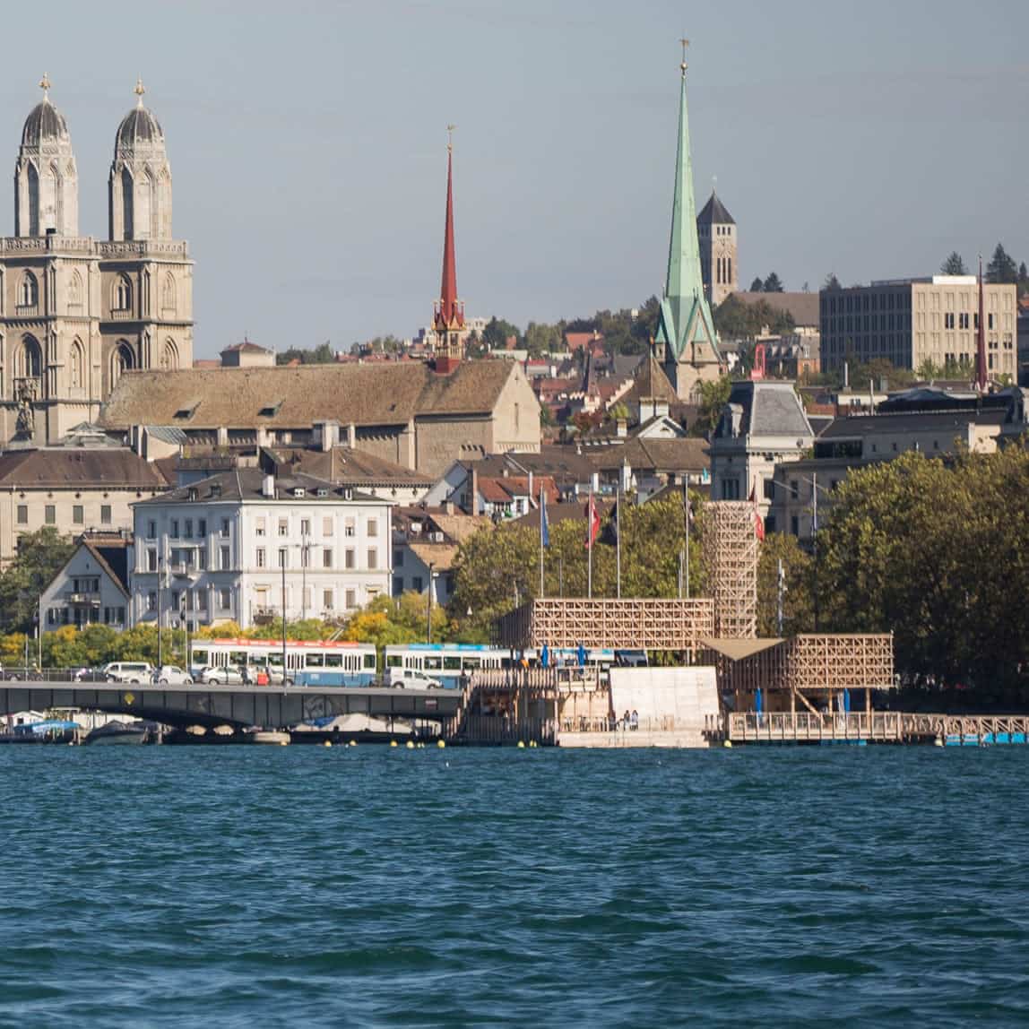 Blick auf die Uferpromenade vom Zürichsee mit historischen Gebäuden und Kirchtürmen mit Zifferblättern, umgeben von Bäumen.