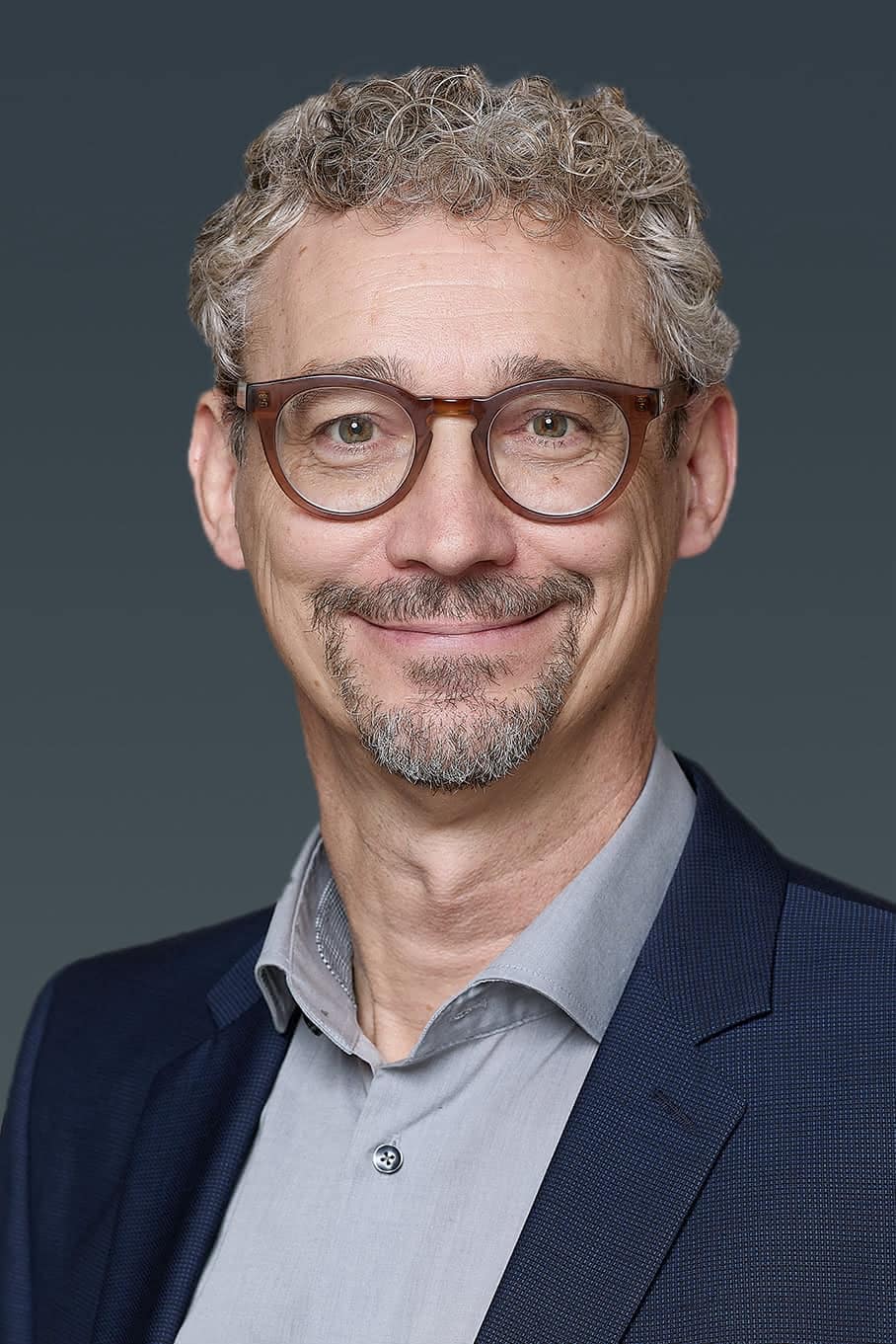 Man with curly hair, glasses, and a goatee wearing a dark suit and light shirt, smiling in front of a gray background.